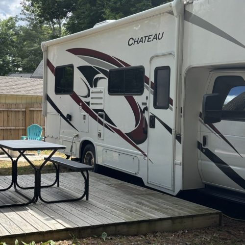 White RV with red/black swoosh graphics parked beside a wooden deck, picnic table and turquoise chair visible outside. The word 'CHATEAU' is on the side.