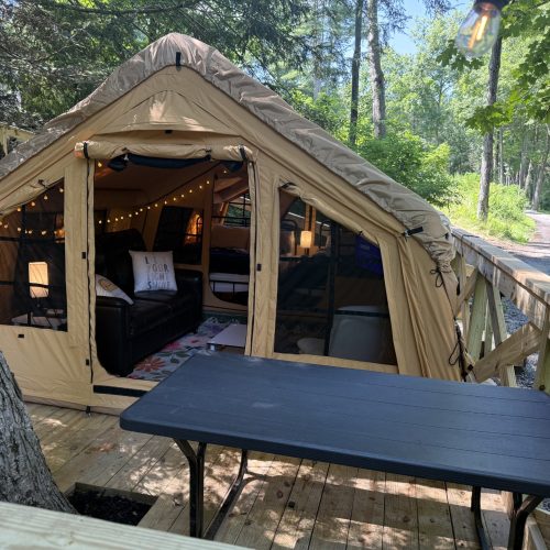 Beige canvas camping tent set up on a wooden deck in a forest with string lights inside and a black sofa visible.