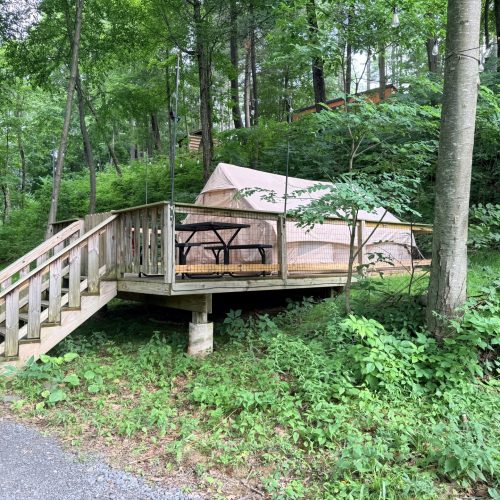 Beige camping tent pitched on a raised wooden deck surrounded by trees in a forest, with a black picnic table nearby and string lights above.