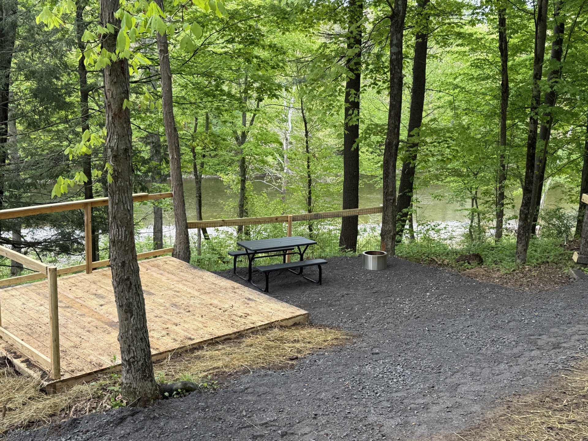Wooden deck in a forest, overlooking a river, with a black picnic table and metal fire pit nearby.