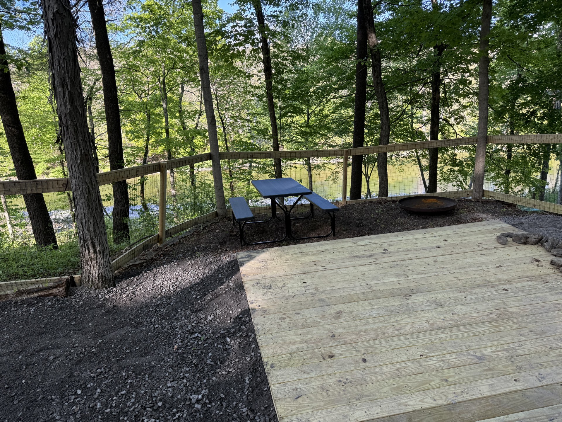Wooden deck with a blue picnic table and benches, overlooking a forest and a wire fence; a fire pit sits nearby.