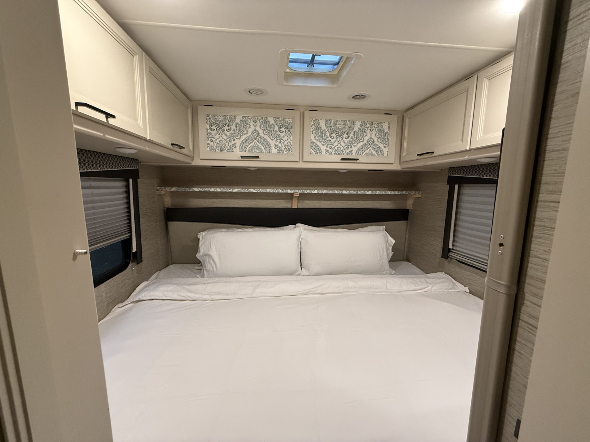 Cozy RV bedroom with a big white bed, overhead white cabinets, and decorative cabinet doors above the headboard. A small skylight and two side windows with blinds provide light and privacy.