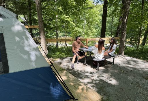 Group of three people sits at a metal table under trees in a campground, with a blue tent on the left and a wooden platform nearby on a sunny day.