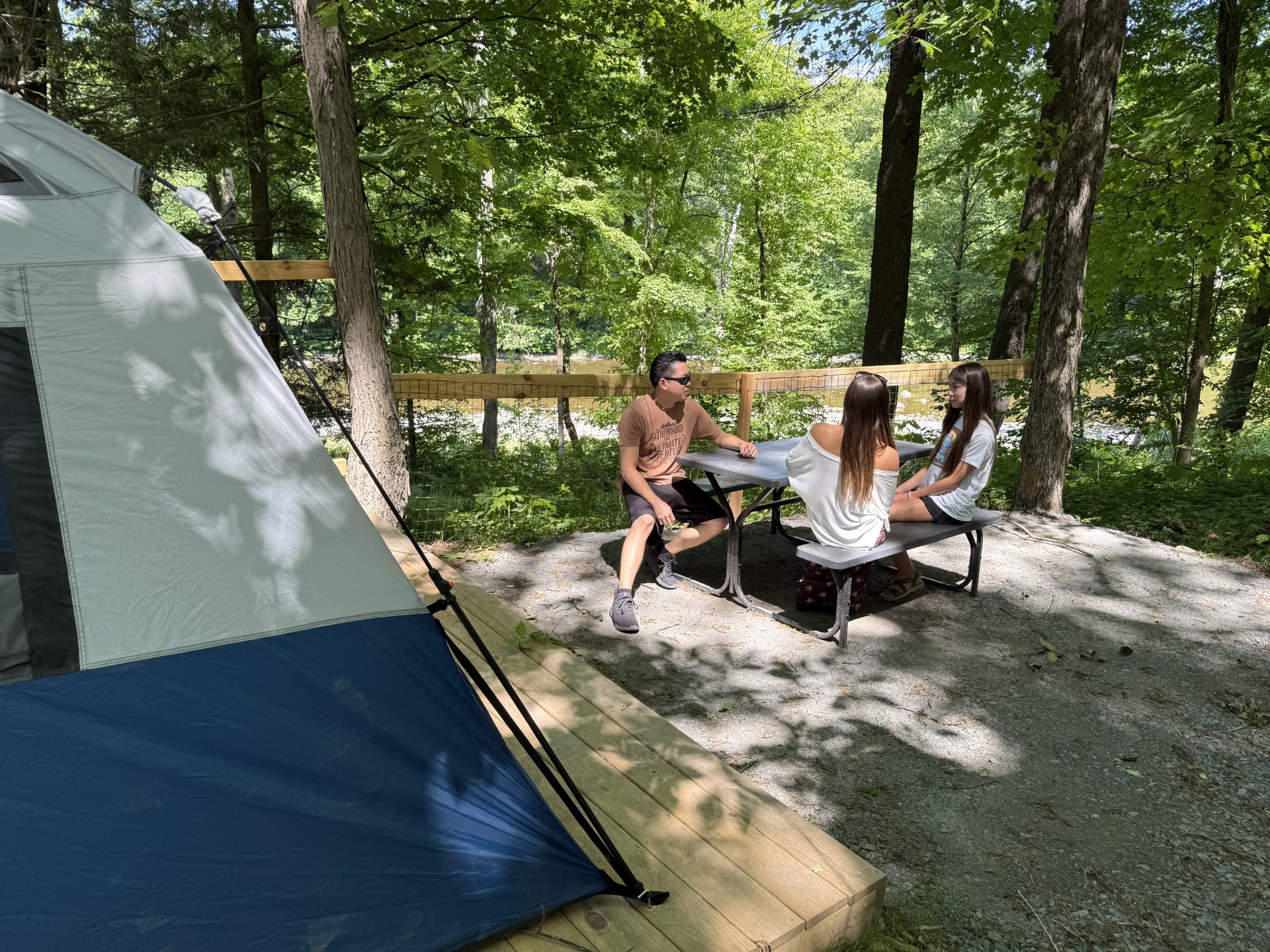 Group of three people sits at a metal table under trees in a campground, with a blue tent on the left and a wooden platform nearby on a sunny day.