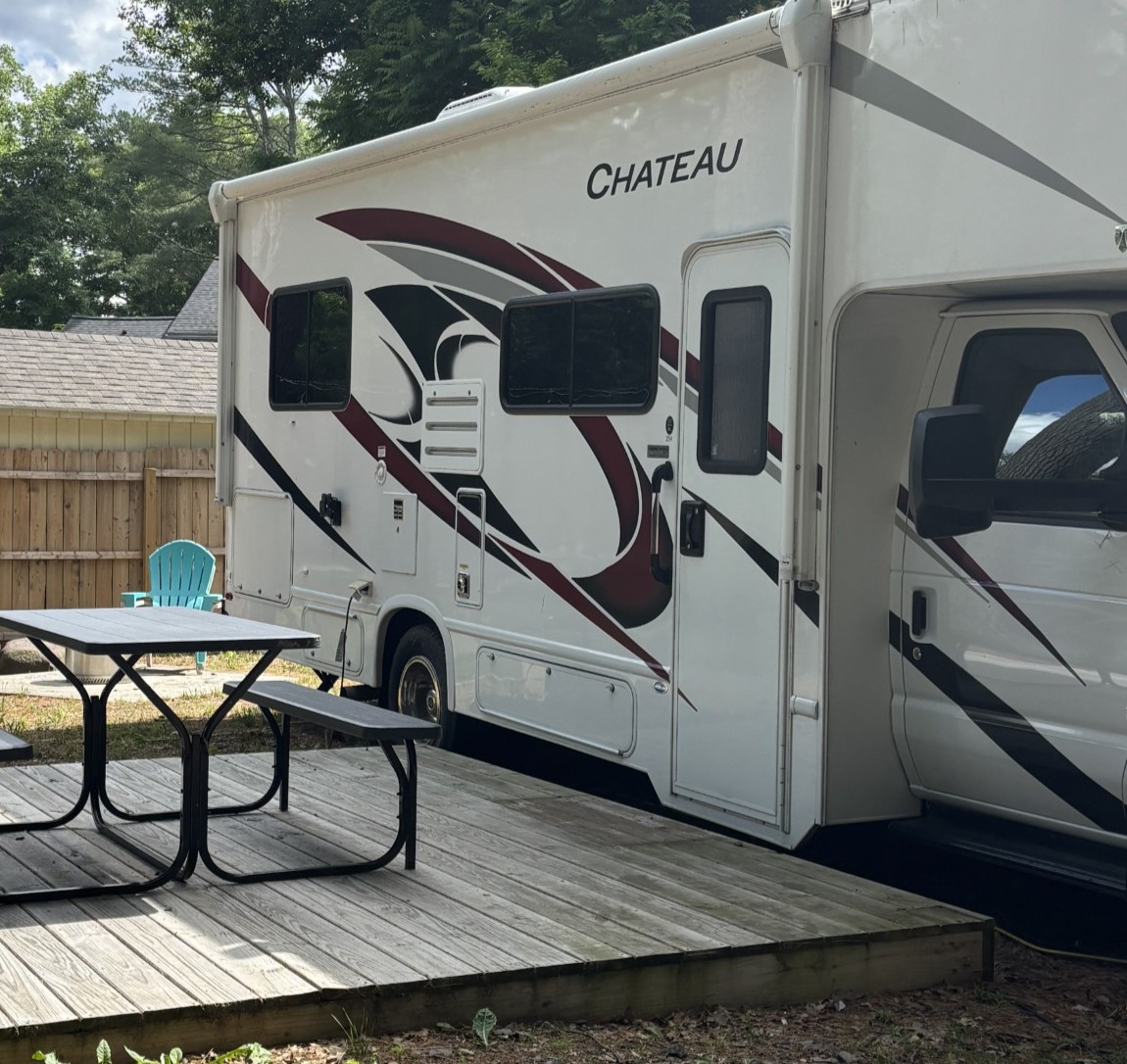White RV with red/black swoosh graphics parked beside a wooden deck, picnic table and turquoise chair visible outside. The word 'CHATEAU' is on the side.
