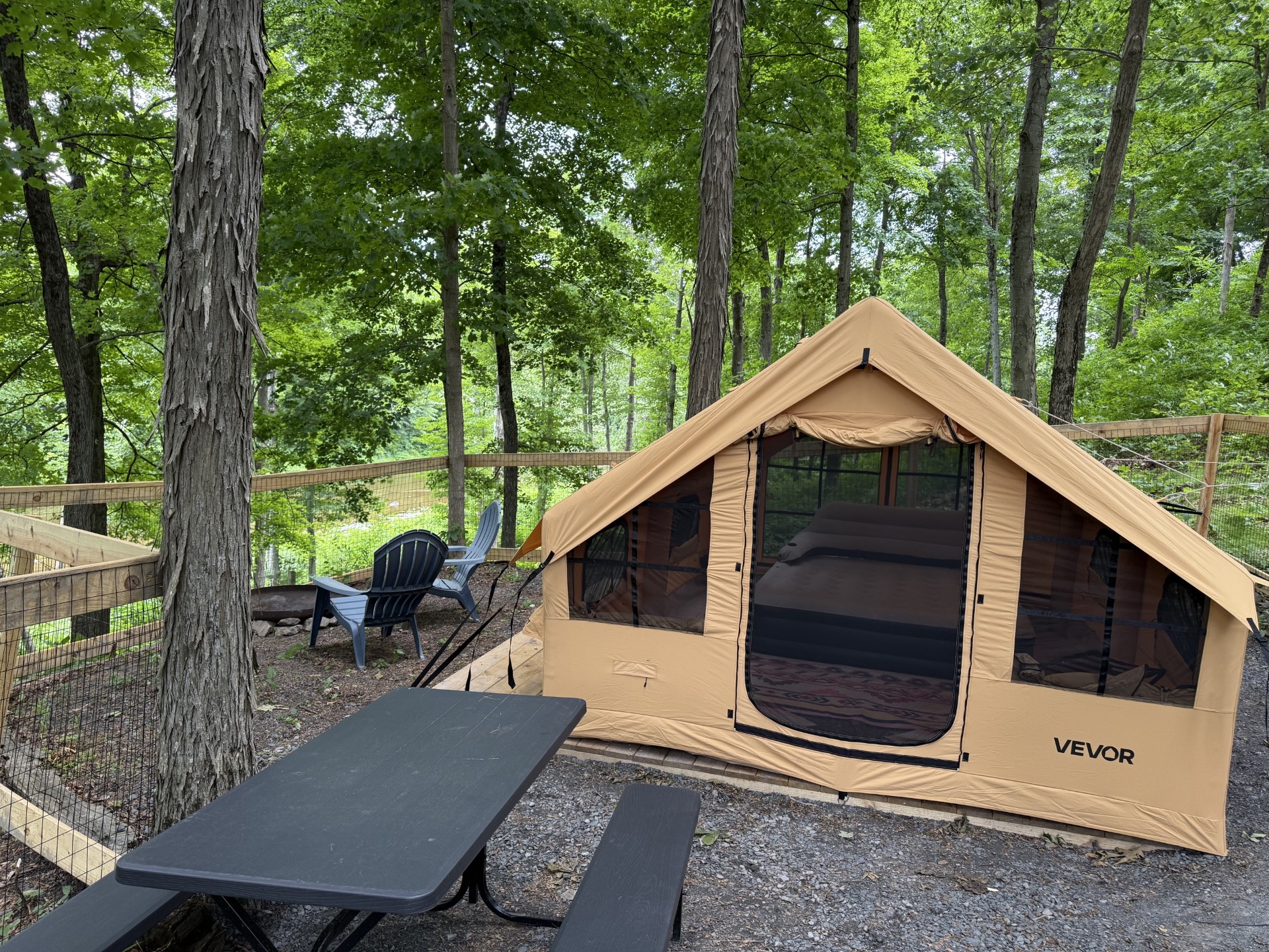 Beige camping tent pitched on a wooden deck in a forested campsite with a fence and plastic chairs nearby.