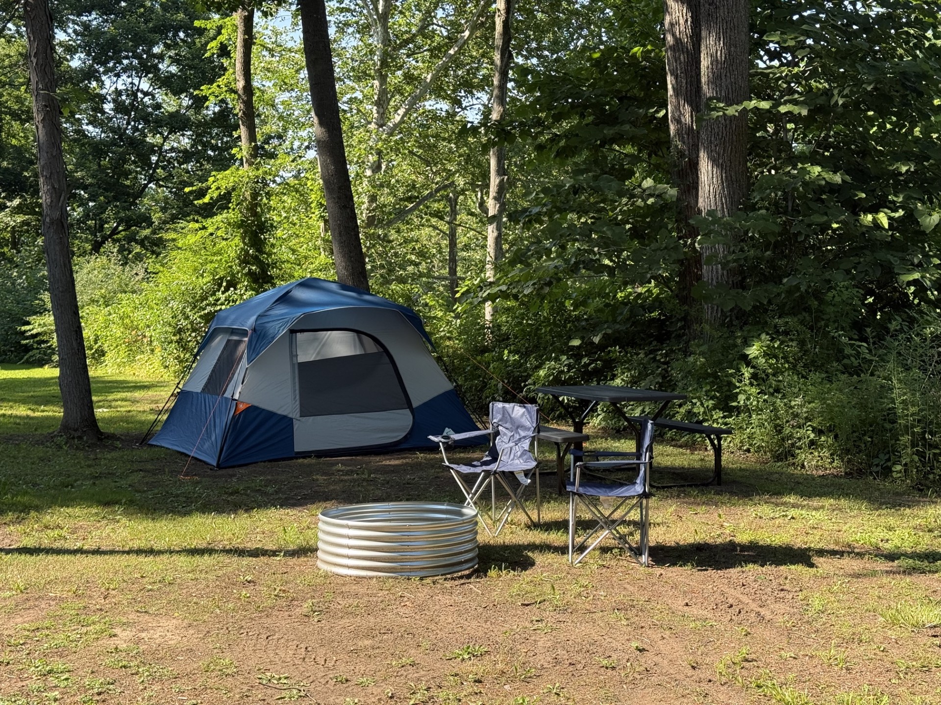Blue and gray camping tent set up on grassy ground in a forested campsite, with folding chairs and a table nearby and a coiled water container in the foreground.