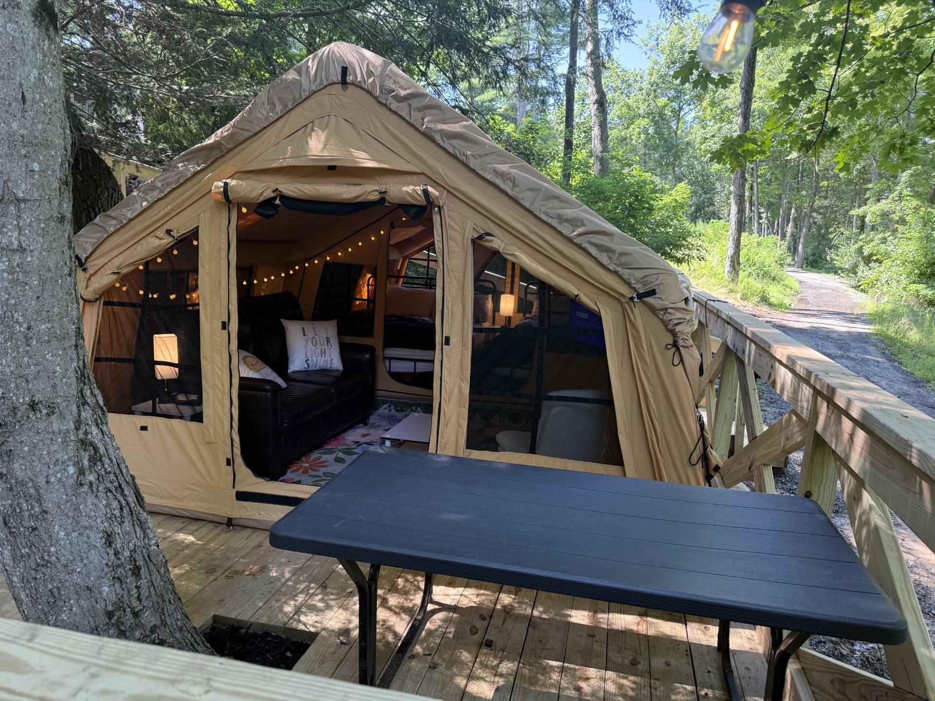 Beige canvas camping tent set up on a wooden deck in a forest with string lights inside and a black sofa visible.